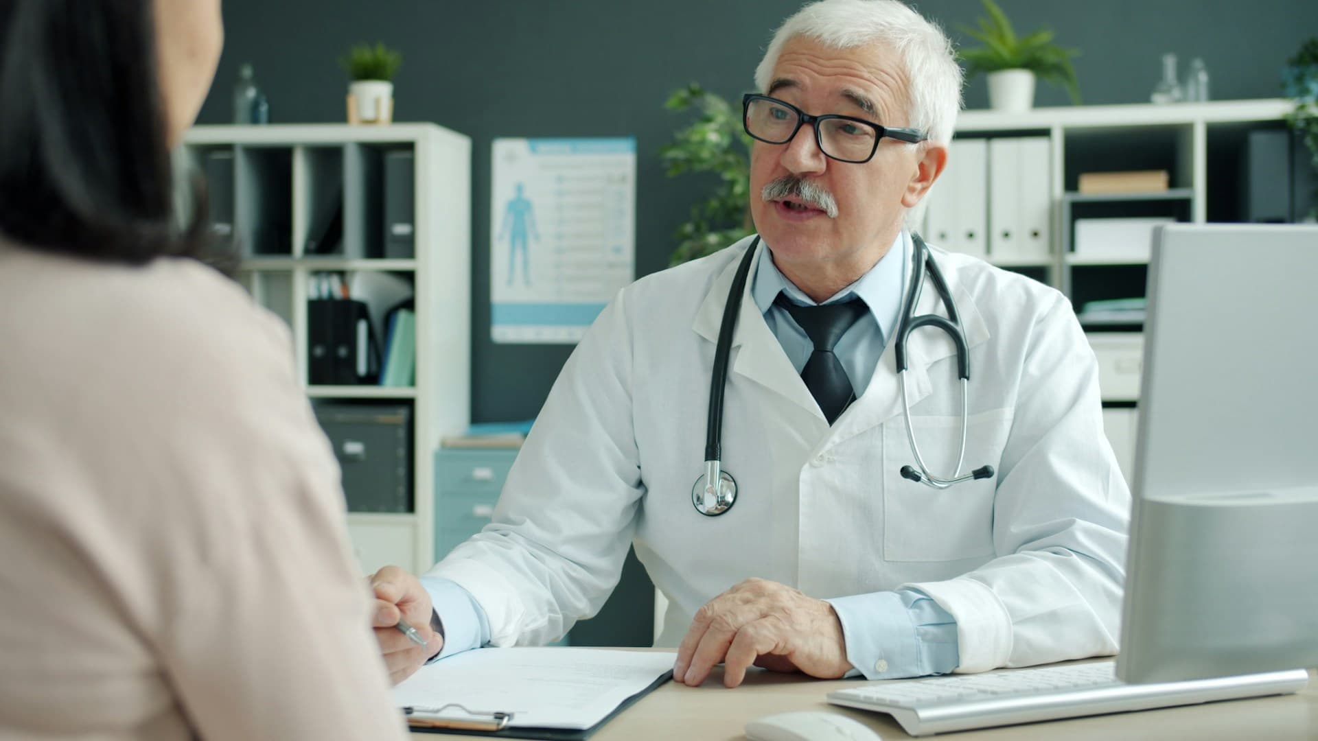 An older male doctor talking to a female patient with a pen and pad in hand